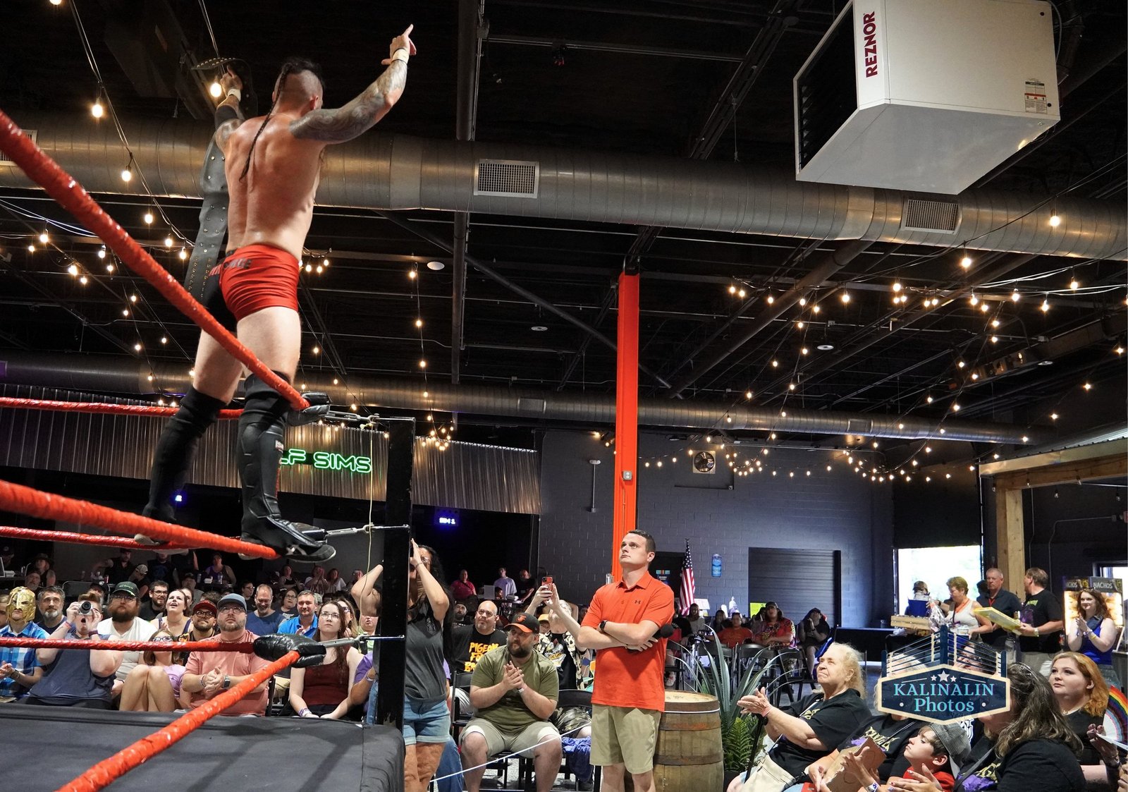 A wrestler in red trunks stands on the ring ropes and points to the crowd at an indoor SlamFest Live show